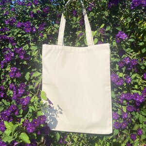May include: A plain white canvas tote bag with two handles hanging from a bush of purple flowers.