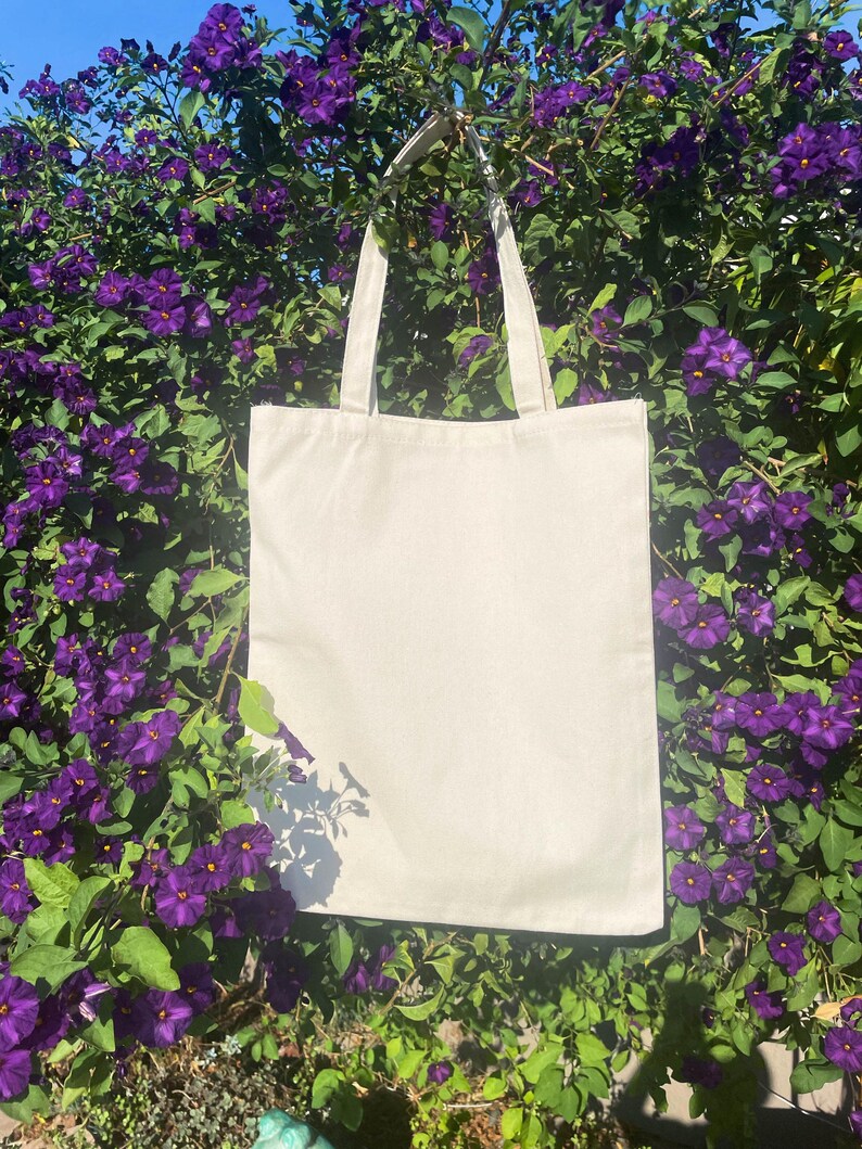 May include: A plain white canvas tote bag with two handles hanging from a bush of purple flowers.