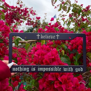 以下が含まれることがあります： 黒色の金属製のナンバープレートフレームに、「believe」と「nothing is impossible with god」という銀色の文字が書かれています。フレームはピンクのブーゲンビリアの花に囲まれています。
