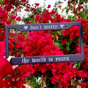 May include: Black license plate frame with the text "don't worry ♥ the world is yours" in a gothic font. The frame is surrounded by pink bougainvillea flowers.