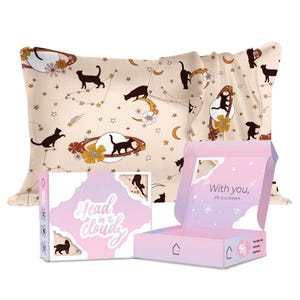 Op de afbeelding: Een roze en paarse doos met de tekst "Head in the clouds" en een kattenillustratie. De doos is open en laat een beige satijnen kussensloop zien met een zwart katten- en sterrenpatroon. De tekst "With you, life is a dream" staat op de doos gedrukt.