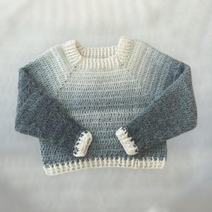 Może przedstawiać: Szydełkowy sweter w odcieniach bieli, szarości i ciemnoszarości. Rękawy i mankiety są ciemnoszare, a korpus przechodzi od bieli do szarości. Mankiety mają mały czarno-biały detal.