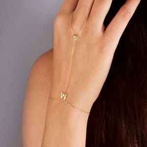 May include: Gold chain bracelet with a green gemstone and a letter 'W' charm. The bracelet is designed to be worn on the wrist and finger.