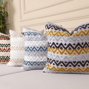 May include: Five decorative throw pillows with a geometric pattern in shades of gray, white, blue, green, and orange. The pillows are arranged on a white couch.