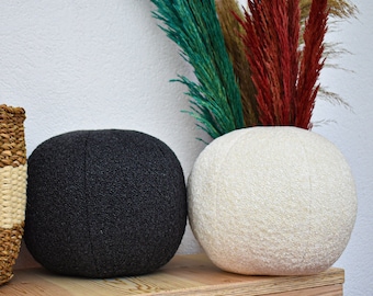 May include: Two round poufs, one black and one white, sit on a wooden table. A bouquet of dried pampas grass with green and red plumes is in the background.