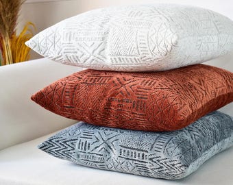 May include: Three decorative throw pillows with geometric patterns. The top pillow is white with a subtle embossed design. The middle pillow is rust-colored with a raised geometric pattern. The bottom pillow is gray with a geometric pattern.