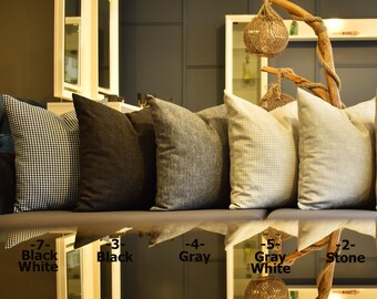 May include: A couch with eight throw pillows in various colors and patterns, including black and white checkered, black, gray, and brown. The pillows are arranged in a row on the couch, with a wooden branch and a hanging light fixture above them. The couch is in front of a wall with a shelf and a mirror.