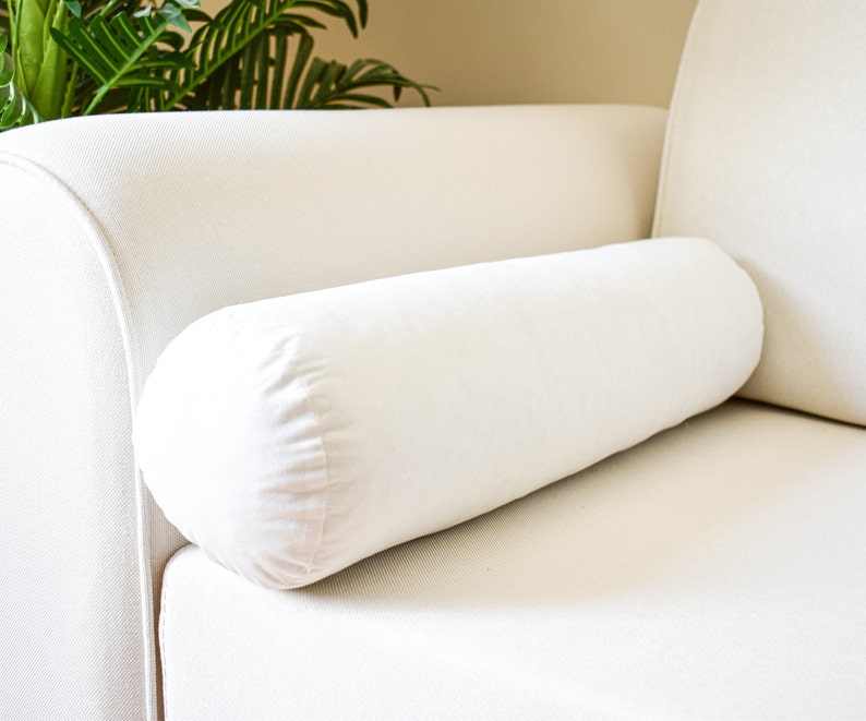 May include: A white cylindrical pillow resting on a white couch. The pillow is made of a soft, plush fabric.