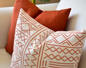 Op de afbeelding: Twee vierkante sierkussens. Eén kussen is effen gebrand oranje. Het andere kussen is wit met een geometrisch patroon in gebrand oranje, bestaande uit horizontale lijnen, zigzaglijnen en ruitvormen op de voorzijde van de stof.