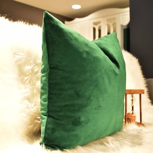 May include: A plush, emerald green throw pillow with a soft, velvety texture. The pillow is resting on a white faux fur surface.