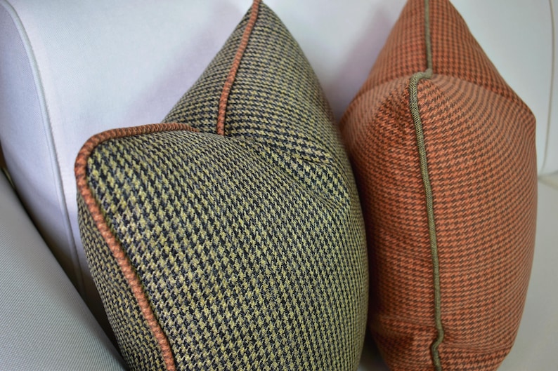 May include: Two decorative throw pillows with a houndstooth pattern. One pillow is olive green and black, the other is orange and black. Both pillows have a contrasting trim.