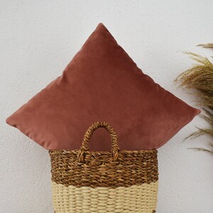 May include: A dusty rose velvet pillow sits on top of a woven wicker basket with a brown and cream pattern.