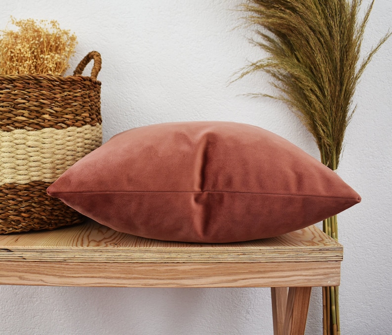 May include: A soft, dusty rose velvet pillow resting on a wooden shelf. The pillow is a rectangular shape with a smooth, plush surface.
