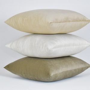 May include: Three decorative throw pillows stacked on top of each other. The top pillow is a light beige color, the middle pillow is a white color, and the bottom pillow is a light brown color.