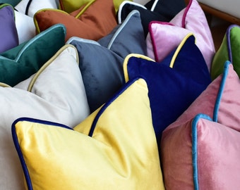 Könnte beinhalten: Eine Sammlung dekorativer Kissen in verschiedenen Farben und Stoffen, darunter Samt, mit kontrastierenden Paspeln. Die Kissen sind in einem Stapel angeordnet und zeigen eine Vielzahl von Texturen und Mustern.
