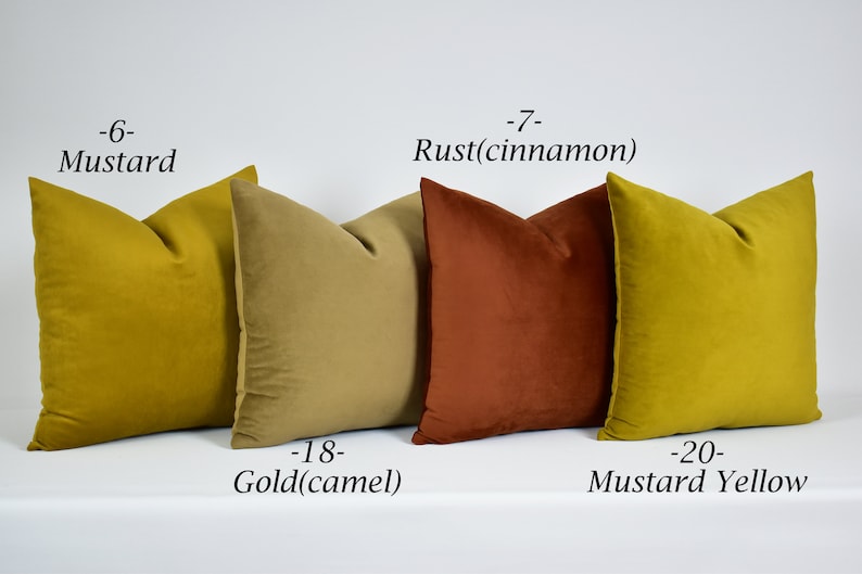 May include: Four decorative throw pillows in various shades of yellow, gold, and rust. The pillows are arranged in a row on a white surface. The pillows are labeled with their colors: Mustard, Rust (cinnamon), Gold (camel), and Mustard Yellow.