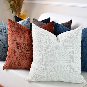 May include: A set of decorative throw pillows with geometric patterns in various colours, including white, grey, blue, and orange. The pillows are arranged on a white sofa.