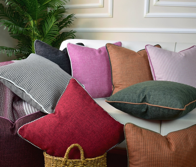May include: A collection of decorative throw pillows in various colors and patterns, including black and white checkered, pink, orange, green, and brown. The pillows are arranged on a white couch.