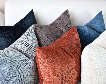 May include: A collection of seven decorative throw pillows with geometric patterns in shades of blue, gray, white, and orange. The pillows are arranged on a white couch.