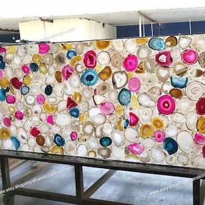 May include: A large, rectangular slab of agate with a white background and colorful agate slices. The agate slices are in various shapes and sizes, and they are arranged in a random pattern. The colors of the agate slices include pink, blue, yellow, and brown. The slab is on a metal stand.