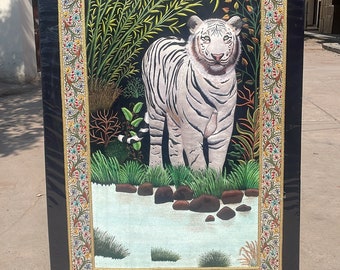 Puede incluir: Una obra de arte de bordado enmarcada que representa un tigre blanco de pie en un entorno de jungla verde exuberante. El tigre está rodeado de follaje y rocas, y el fondo es un cielo azul oscuro. El marco es negro con un borde dorado y diseños florales intrincados.