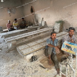 May include: A workshop with several men working on a project. They are using a scale to weigh materials and there are several wooden frames with white tiles on them. The workshop is dusty and has a concrete floor.