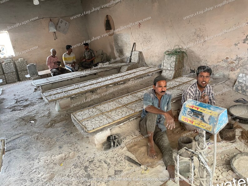 May include: A workshop with several men working on a project. They are using a scale to weigh materials and there are several wooden frames with white tiles on them. The workshop is dusty and has a concrete floor.
