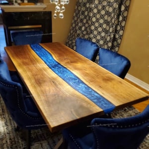 May include: A large wooden dining table with a blue epoxy resin river running down the center. The table is surrounded by six blue velvet chairs with black metal legs.