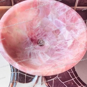 Pode incluir: Uma pia redonda em forma de ta&ccedil;a feita de pedra de quartzo rosa transl&uacute;cida. A bacia apresenta uma superf&iacute;cie interior polida e lisa com fraturas cristalinas naturais vis&iacute;veis e uma abertura de drenagem circular no fundo.