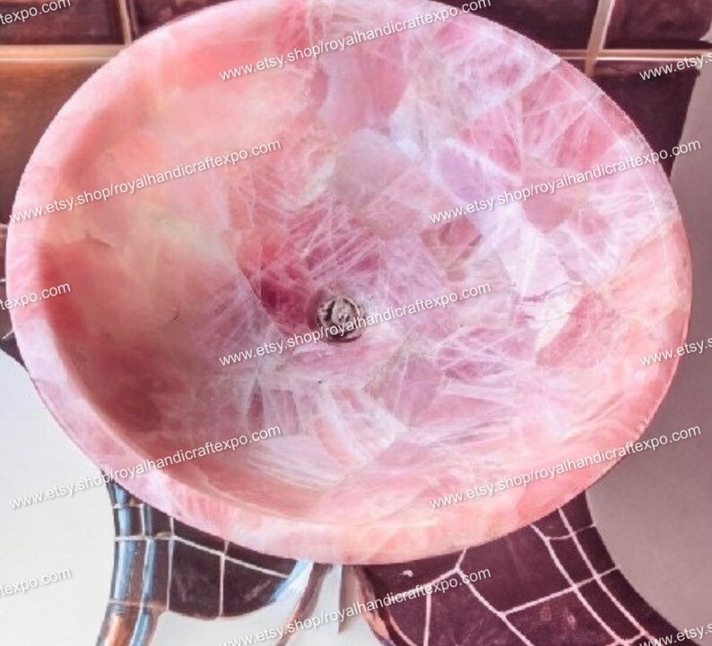 Pode incluir: Uma pia redonda em forma de ta&ccedil;a feita de pedra de quartzo rosa transl&uacute;cida. A bacia apresenta uma superf&iacute;cie interior polida e lisa com fraturas cristalinas naturais vis&iacute;veis e uma abertura de drenagem circular no fundo.