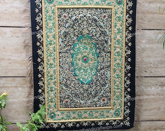 Puede incluir: Un tapiz de pared de terciopelo negro con intrincados bordados dorados y verdes. El diseño presenta un motivo floral central con una joya roja en el centro. El tapiz de pared está rodeado por un borde de bordado dorado y verde.