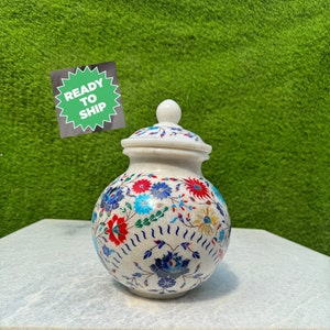 May include: A round white stone jar with a matching lid, featuring intricate floral inlay work in vibrant red, blue, and green colours. The surface is decorated with detailed flower patterns and curved lines. A green sticker reads READY TO SHIP.