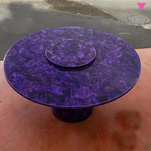 Op de afbeelding: Een ronde, paarse tafel met een bijpassende kleinere, cirkelvormige inzet in het midden. De tafel heeft een voetstuk en een gemarmerd patroon. De diameter van de tafel is ongeveer 122 cm.