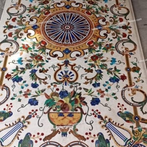 May include: A white marble floor tile with a colorful mosaic design. The design features a central circular motif with floral patterns and birds. The tile is surrounded by a decorative border.