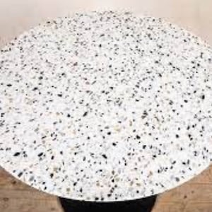 Op de afbeelding: Een ronde terrazzo tafelblad met een witte achtergrond en zwarte en bruine vlekken.