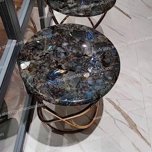 May include: Two round side tables with a dark, speckled tabletop and a gold-colored, curved metal base. The tables are made of a dark, speckled stone with blue and green hues.