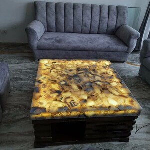 May include: A coffee table with a unique agate stone top. The top is backlit, illuminating the intricate patterns of the stone in shades of brown, yellow, and white. The table has a dark wood base.