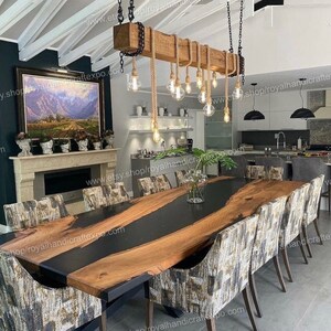 May include: A long wooden dining table with a black epoxy river running through the center. The table is surrounded by chairs with patterned fabric upholstery. A rustic wooden chandelier with rope accents hangs above the table.