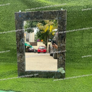 May include: A rectangular mirror with a dark green, brown, and blue mosaic frame. The mirror is reflecting a view of a street with cars and trees.