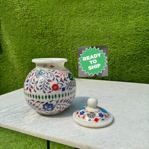 May include: White marble decorative jar with a floral design in blue, red, and green. The jar has a lid with a similar floral design. The jar is on a white surface.