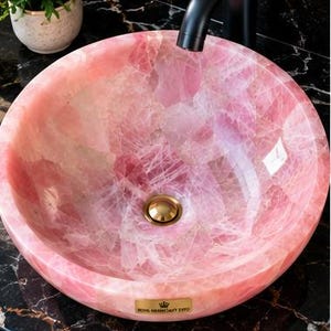 Pode incluir: Uma cuba de apoio redonda feita de pedra de quartzo rosa transl&uacute;cido com acabamento polido. A bacia apresenta uma v&aacute;lvula de escoamento em tom dourado ao centro e uma pequena placa met&aacute;lica retangular com um log&oacute;tipo de coroa e texto na borda.
