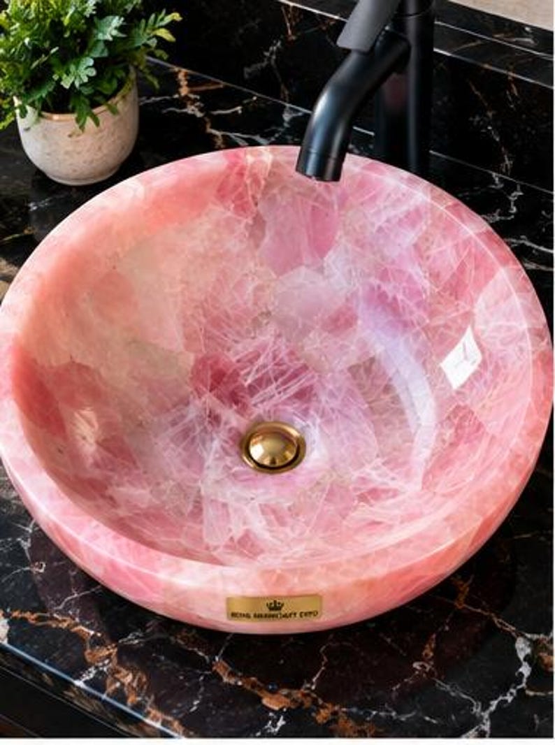 Pode incluir: Uma cuba de apoio redonda feita de pedra de quartzo rosa transl&uacute;cido com acabamento polido. A bacia apresenta uma v&aacute;lvula de escoamento em tom dourado ao centro e uma pequena placa met&aacute;lica retangular com um log&oacute;tipo de coroa e texto na borda.