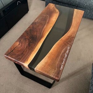May include: A wooden coffee table with a black epoxy resin river flowing through the center. The table has a black metal base and is made by Royal Handicraft Exports.