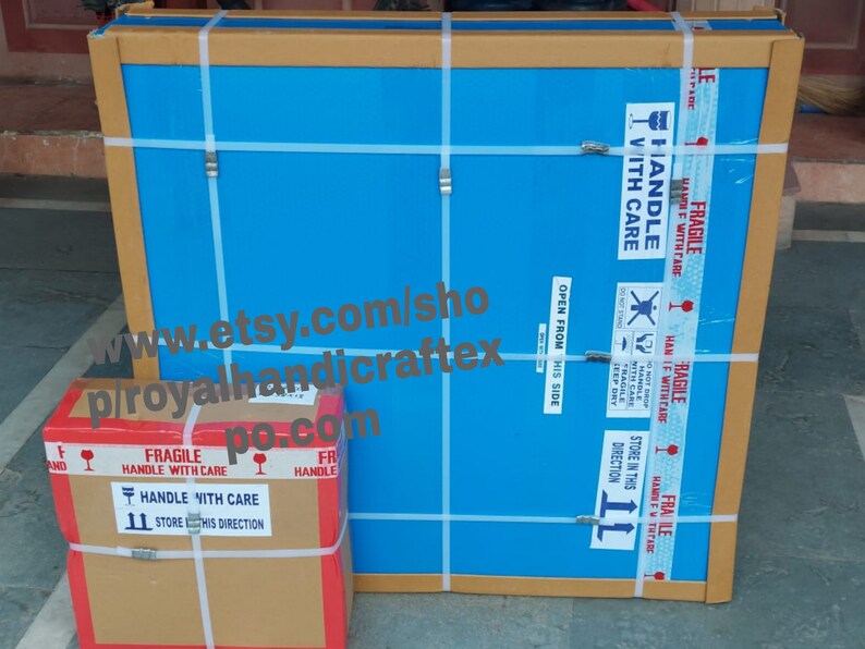 May include: Two cardboard boxes with blue and brown tape. The larger box has a blue surface with the text "HANDLE WITH CARE" and "OPEN FROM THIS SIDE". The smaller box has a brown surface with the text "FRAGILE HANDLE WITH CARE" and "STORE IN THIS DIRECTION". The text "www.etsy.com/shop/p/royalhandicraftex.po.com" is visible on the boxes.