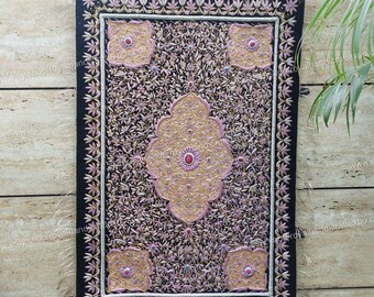 Puede incluir: Un tapiz de terciopelo negro con intrincados bordados en hilo dorado y rosa. El diseño presenta un motivo central de una flor estilizada con patrones florales más pequeños que la rodean. El tapiz tiene un borde con flecos.