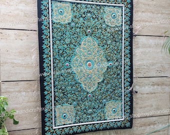 Puede incluir: Un tapiz de pared rectangular negro con un intrincado diseño floral turquesa y dorado. El motivo central es un patrón floral estilizado, rodeado por un borde turquesa y detalles dorados. La pieza está adornada con pequeños adornos rojos y tiene un borde con flecos.
