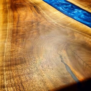 May include: Close-up of a wooden table top with a blue epoxy resin river running down the center. The wood has a rich brown color and a natural grain pattern.