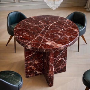 May include: A circular dining table with a red marble top, supported by a distinctive pedestal base. The table is encircled by four chairs with dark green leather seats and wooden legs. A crystal chandelier hangs above. The table is approximately 120 cm in diameter.