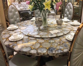 May include: A round dining table with a unique stone mosaic top, featuring a central lazy susan. The table is surrounded by upholstered chairs with gold trim. A vase of lilies and teacups complete the elegant setting. The table is approximately 60 inches in diameter.