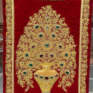 May include: A red velvet wall hanging with a gold embroidered floral design. The flowers are arranged in a vase with a gold and green design.